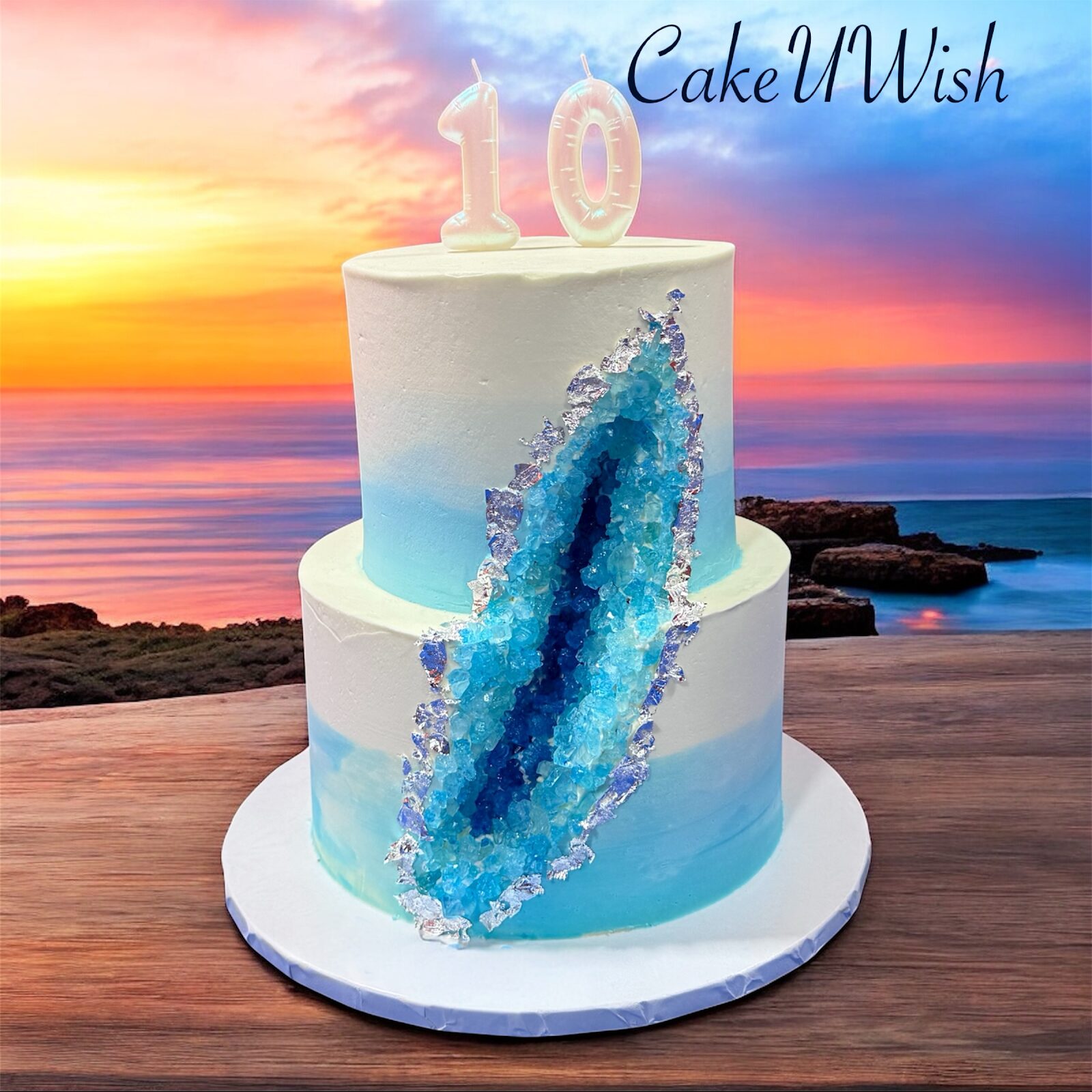 Teal and white two-tier geode drip cake with a gold '10' topper, on a beach at sunset